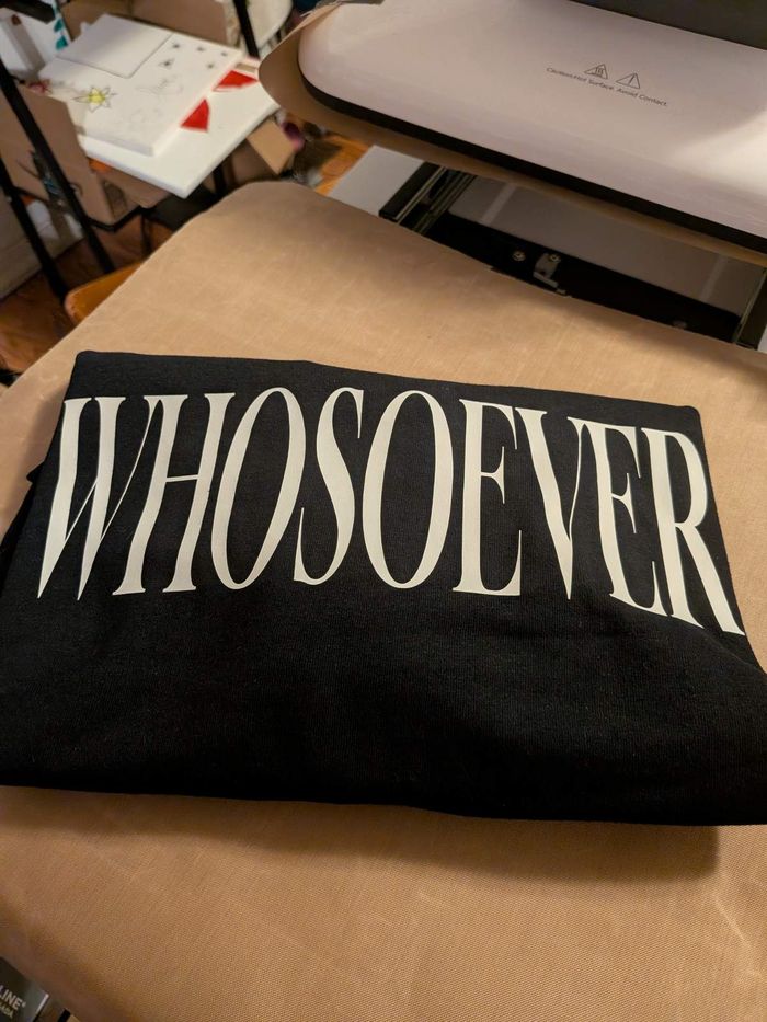 A folded black WHOSOEVER T-shirt resting on the press mat, with the heat press open behind it.