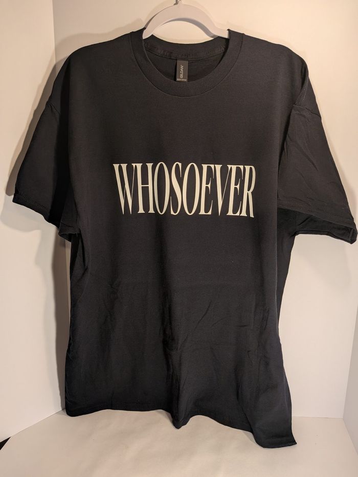 A black WHOSOEVER T-shirt hanging on a hanger against a plain wall.